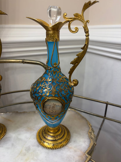 Stunning antique 19th-century French Napoleon III-style blue opaline glass ewer jugs pair adorned with elaborate gilt ormolu (gold-plated bronze) mounts, scrollwork, and scenic medallions! Circa 1836-1850