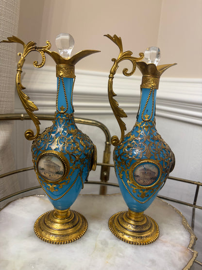 Stunning antique 19th-century French Napoleon III-style blue opaline glass ewer jugs pair adorned with elaborate gilt ormolu (gold-plated bronze) mounts, scrollwork, and scenic medallions! Circa 1836-1850